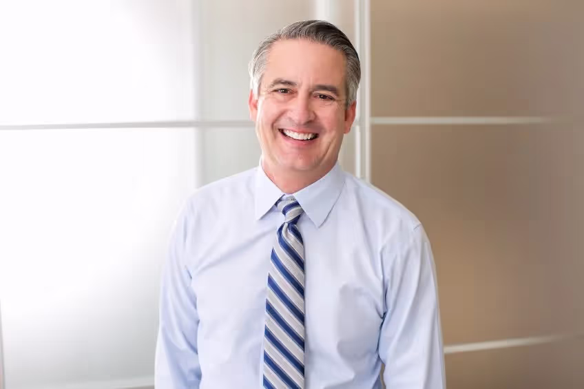 Smiling middle-aged man wearing a light blue dress shirt and striped tie standing indoors against a blurred background.