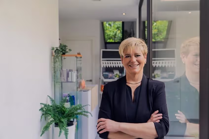 Smiling woman with short blonde hair standing with arms crossed in a modern office space with plants and glass reflections.