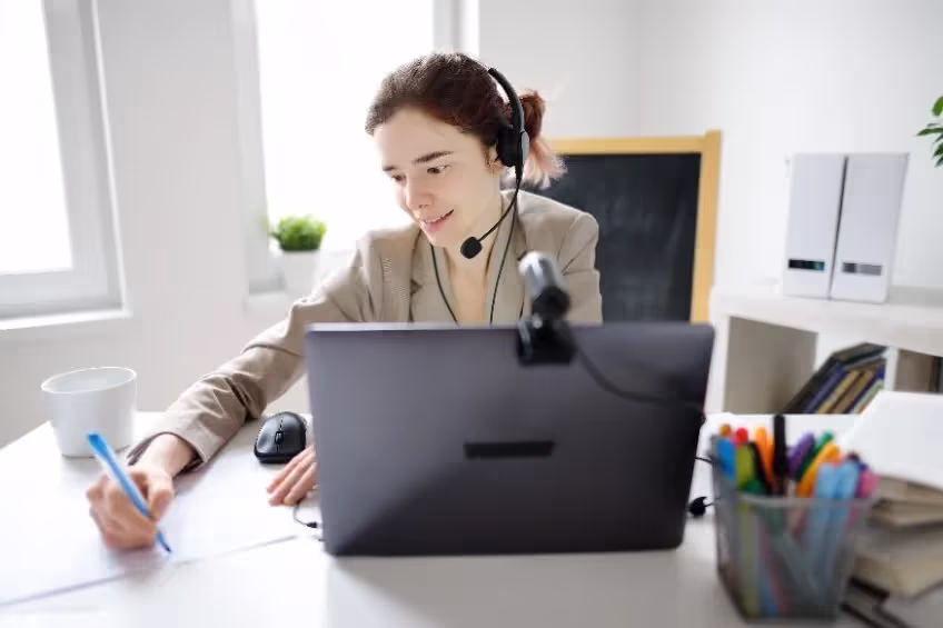Young woman wearing headset writing notes while working on a laptop in a bright office.